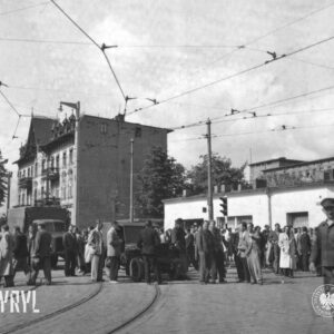 duża grupa ludzi, przeważnie mężczyzn zgromadzona na skrzyżowaniu ulic, pomiędzy ludźmi samochody, w głębi kilkupiętrowa kamienica
