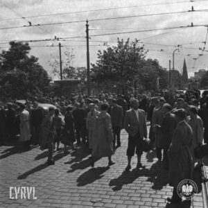 tłum ludzi na skrzyżowaniu ulic, pośrodku słupy i linie trakcji tramwajowej, w głębi drzewa