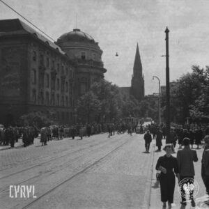tłum ludzi idący lub stojący na chodniku i ulicy, z lewej okazały gmach i wysoka strzelista wieża w otoczeniu drzew