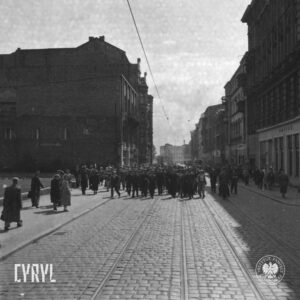 tłum ludzi idzie całą szerokością brukowanej ulicy, w pierwszych rzędach mężczyźni w roboczych kombinezonach, po obu stronach ulicy kamienice