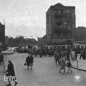 brukowany plac, na nim skupiska ludzi, w centrum między ludźmi trawaj, w głębi samotna kamienica