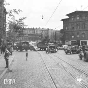 pośrodku brukowanej ulicy chaotycznie stojące samochody osobowe i ciężarowe, na chodnikach grupy ludzi i pojedynczych osób, część stoi, część idzie, w głębi kilkupiętrowe kamienice