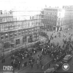 grupy ludzi stojące na ulicy, pośrodku czołg, z lewej kamienica otoczona drewnianym rusztowaniem, z prawej narożna kamienica