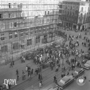 grupy ludzi stojące na ulicy, pośrodku czołg, z lewej kamienica otoczona drewnianym rusztowaniem, z prawej narożna kamienica