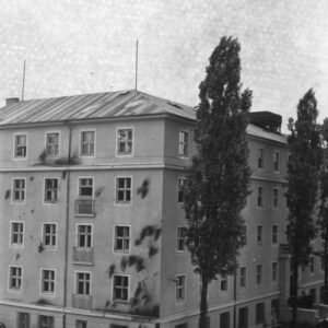 pięciokondygnacyjny budynek, na jego elewacji ciemne ślady po obrzuceniu budynku palącymi się butelkami z benzyną, powybijane szyby, z prawej wysokie drzewa, chodnikiem spacerują ludzie