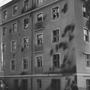 pięciokondygnacyjny budynek, na jego elewacji ciemne ślady po obrzuceniu budynku palącymi się butelkami z benzyną, powybijane szyby, przed budynkiem kilku mężczyzn