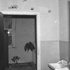 fragment pomieszczenia biurowego, na ścianie z prawej wisi umywalka, na wprost otwarte drzwi, za nimi widać drugie pomieszczenie z oknem, w oknie podarte firanki, na podłodze roślina w doniczce, nad drzwiami, na ścianie widoczne ślady po ostrzale