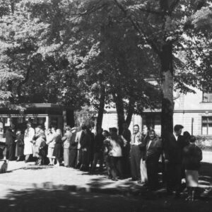 ludzie stojący w kolejce do małej budki handlowej, stojącej w otoczeniu drzew, z prawej słup ogłoszeniowy, w głębi budynek