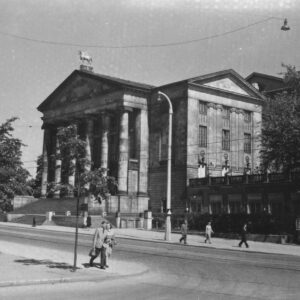 tramwaje jadące ulicą, w głębi okazały gmach z portykiem i kolumnami, pojedynczy przechodnie