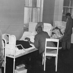 dwa łóżka szpitalne stoją wzdłuż ściany, na nich leżą chorzy, przy jednym z łóżek siedzi kobieta na krześle, w głębi przy drugim łóżku stoi mężczyzna, na pierwszym planie, z lewej stoi mężczyzna w jasnym prochowcu