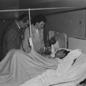 mężczyzna leży na szpitalnym łóżku, przy łóżku chorego stoją kobieta i mężczyzna