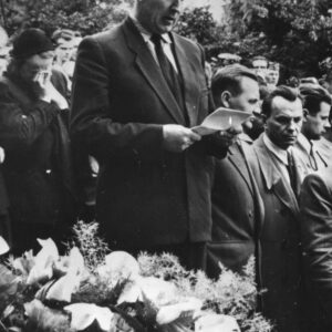 mężczyzna stoi z kartką w rękach i odczytuje tekst, za nim stoi kilkanaście osób, na pierwszym planie kwiaty