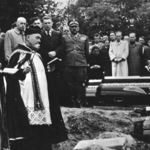 z prawej trzy trumny, z lewej ksiądz stoi nad jedną z trumien, w głębi kilkanaście osób