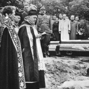 z prawej trzy trumny, z lewej ksiądz stoi nad jedną z trumien, w głębi duża grupa osób