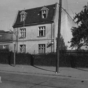 niewielki trzykondygnacyjny budynek za ogrodzeniem z siatki, wzdłuż płotu żywopłot