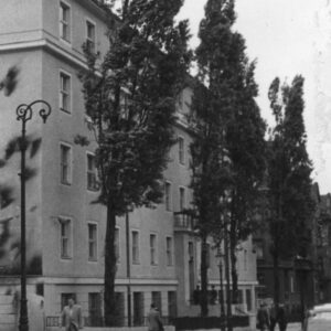 pięciokondygnacyjny budynek, na jego elewacji ciemne ślady po obrzuceniu budynku palącymi się butelkami z benzyną, przed budynkiem wysokie drzewa, chodnikiem spacerują ludzie