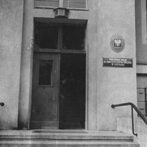 kilka stopni prowadzących do drzwi wejściowych do budynku, prawe skrzydło otwarte, nad wejściem numer, na ścianie metalowa tabliczka z godłem i nazwą urzędu