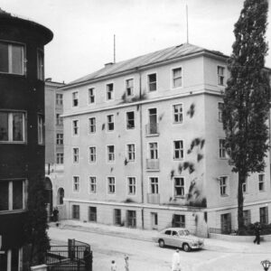pięciokondygnacyjny budynek, na jego elewacji ciemne ślady po obrzuceniu budynku palącymi się butelkami z benzyną, powybijane szyby, przed budynkiem zaparkowany samochód osobowy