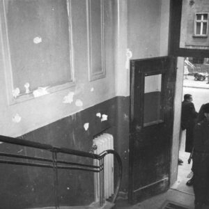 klatka schodowa, na ścianie ślady po ostrzale, u dołu uchylone drzwi na zewnątrz budynku, w drzwiach stoi mężczyzna w mundurze