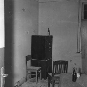 pomieszczenie biurowe z meblami biurowymi, na ścianach ślady po ostrzale, na podłodze gruz, w narożniku pomieszczenia metalowa szafa, na biurku z prawej stoi butelka