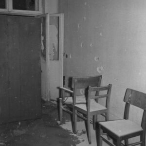 pomieszczenie biurowe, pod ścianą stoją trzy krzesła, na ścianie ślady po ostrzale, z lewej szafa, za nią widoczne otwarte okno balkonowe z wybitymi szybami