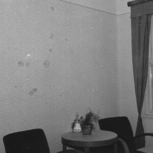 fragment pomieszczenia biurowego, na ścianie widoczne ślady po ostrzale, pod ścianą stolik na nim doniczka z rośliną i karafka, po obu stronach stolika fotele