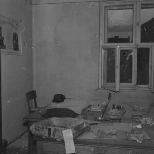 fragment pomieszczenia biurowego, pośrodku na biurkach porozrzucane rzeczy, okno z wybitymi szybami, na ścianie ślady po ostrzale