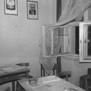 fragment pomieszczenia biurowego, pośrodku stoją biurka i krzesła, na ścianie na wprost wisi godło państwowe i zdjęcie portretowe mężczyzny, z prawej otwarte okno z powybijanymi szybami