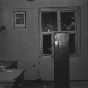 fragment pomieszczenia biurowego, pod ścianą z lewej biurka, na ścianie zdjęcie portretowe mężczyzny w mundurze, na ścianie z prawej widoczne ślady po ostrzale, na wprost okno, pod nim stoi szafa