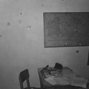 fragment pomieszczenia biurowego, na ścianie wisi mapa województwa poznańskiego, na mapie i wokół niej ślady po ostrzale, pośrodku stoi biurko, na nim rozrzucone papiery, z prawej wieszak z ubraniami