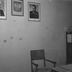 fragment pomieszczenia biurowego, na ścianie pośrodku wisi godło państwowe, po jego obu stronach portrety zdjęcia mężczyzn, na ścianie widoczne ślady po ostrzale, w narożniku z prawej stoi metalowa szafa, na niej przewieszona firanka