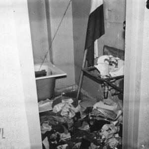 fragment zdemolowanej łazienki, na podłodze porozrzucane rzeczy i śmieci, z prawej rozbita umywalka, przy niej oparta o ścianę polska flaga na drzewcu