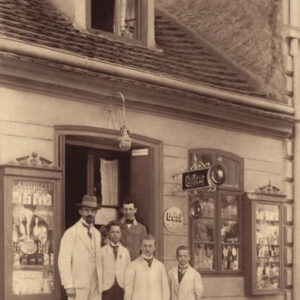 grupa mężczyzn w różnym wieku ubranych w fartuchy fryzjerskie i pozujących do fotografii na tle kamienicy, na której wiszą szyldy