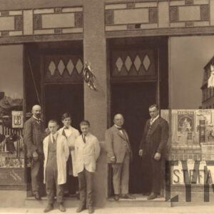 grupa mężczyzn w różnym wieku, kilku z nich nosi fartuchy fryzjerskie, kilku garnitury; wszyscy pozują do fotografii na tle kamienicy, z dużymi, przeszklonymi witrynami reklamowymi