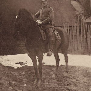 młody mężczyzna w mundurze Wojska Polskiego, siedzący na koniu, przy siodle wisi przytroczona szabla; w tle zabudowa wiejska - chaty i drewniany płot