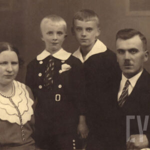 mężczyzna i kobieta w średnim wieku oraz dwójka młodych chłopców pozujący do zdjęcia, wszyscy ubrani są w odświętne, eleganckie stroje; mężczyzna w garnitur, koszulę i krawat, kobieta w suknię udekorowaną żabotem; chłopcy w marynarki i białe koszule 