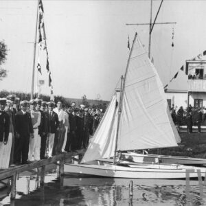 tłum ludzi zebrany na brzegu jeziora i drewnianym pomoście, przy pomoście zacumowane trzy żaglówki, w głębi niewielki drewniany budynek
