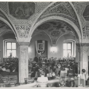 liczna grupa elegancko ubranych ludzi, głównie mężczyzn, część z nich siedzi za stołem prezydialnym, część na krzesłach naprzeciwko stołu; na suficie widoczne bogate freski i zdobienia; na ścianie za plecami mężczyzn wisi duży portret mężczyzny z wąsami