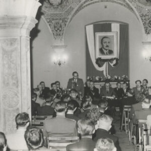 liczna grupa elegancko ubranych ludzi, głównie mężczyzn, część z nich siedzi za stołem prezydialnym, część na krzesłach naprzeciwko stołu; na suficie widoczne bogate freski i zdobienia; na ścianie za plecami mężczyzn wisi duży portret mężczyzny z wąsami