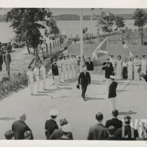 pośrodku kilku mężczyzn w ciemnych ubraniach, część w białych spodniach, w głębi kilkanaście osób w jasnych ubraniach stoi w jednym szeregu, w oddali jezioro