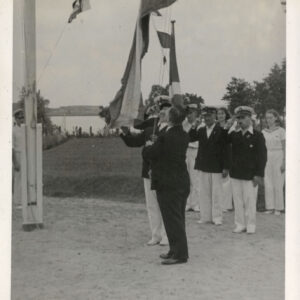 dwóch mężczyzn wciąga flagę na maszt, w głębi, w głębi dwóch mężczyzn w ciemnych marynarkach i jasnych spodniach salutuje, za nimi kilka osób w jasnych ubraniach 