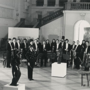 duża sala, kobiety i mężczyźni stoją z instrumentami muzycznymi w dłoniach, elegancko ubrani, przed nimi stoi kobieta z batutą w dłoni, w głębi ozdobne schody