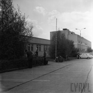 na pierwszym planie kilku młodych mężczyzn z łopatami, kilku kuca przy metalowym koszu na śmieci, w głębi prosty budynek