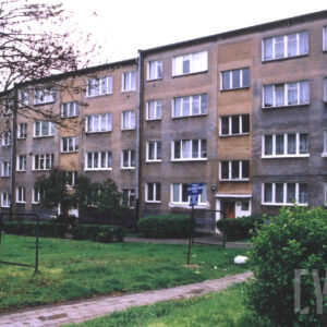 czterokondygnacyjny dom mieszkalny złożony z trzech modułów, każdy moduł z osobną klatką schodową, wzdłuż bloku chodnik osłonięty zielonymi krzakami, na pierwszym planie konstrukcja do wieszania prania