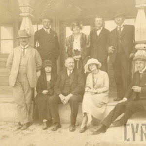 grupa elegancko ubranych osób w różnym wieku, pozujących do fotografii na tle drewnianego domku letniskowego