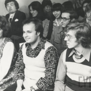 w pierwszym rzędzie siedzą młodzi, kobieta (z lewej) i dwóch mężczyzn, w głębi dalsze rzędy ludzi