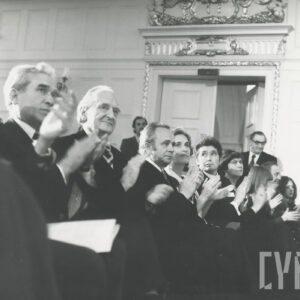 około dziesięciu osób siedzi w jednym rzędzie, wszyscy klaszczą, w głębi ozdobne drzwi