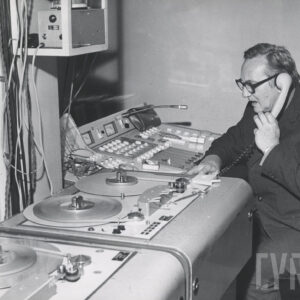 mężczyzna siedzi przy pulpicie radiowym, przy uchu trzyma słuchawkę telefoniczną