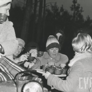 kobieta z prawej trzyma na tacy kilka filiżanek z napojem i częstuje nim kilka osób siedzących na bryczce