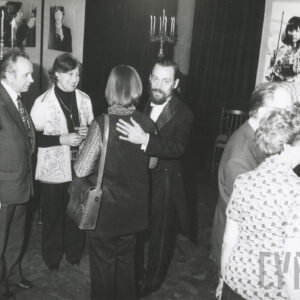 kilka osób zgromadzonych w sali wystawowej rozmawia ze sobą, w głębi zawieszone duże fotografie, między nimi ciemne tkaniny